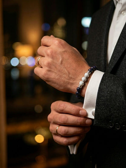 A close-up lifestyle shot of a man's hands adjusting his cufflinks while wearing a dark gray suit jacket, white dress shirt, and a dark blue beaded bracelet on his wrist.