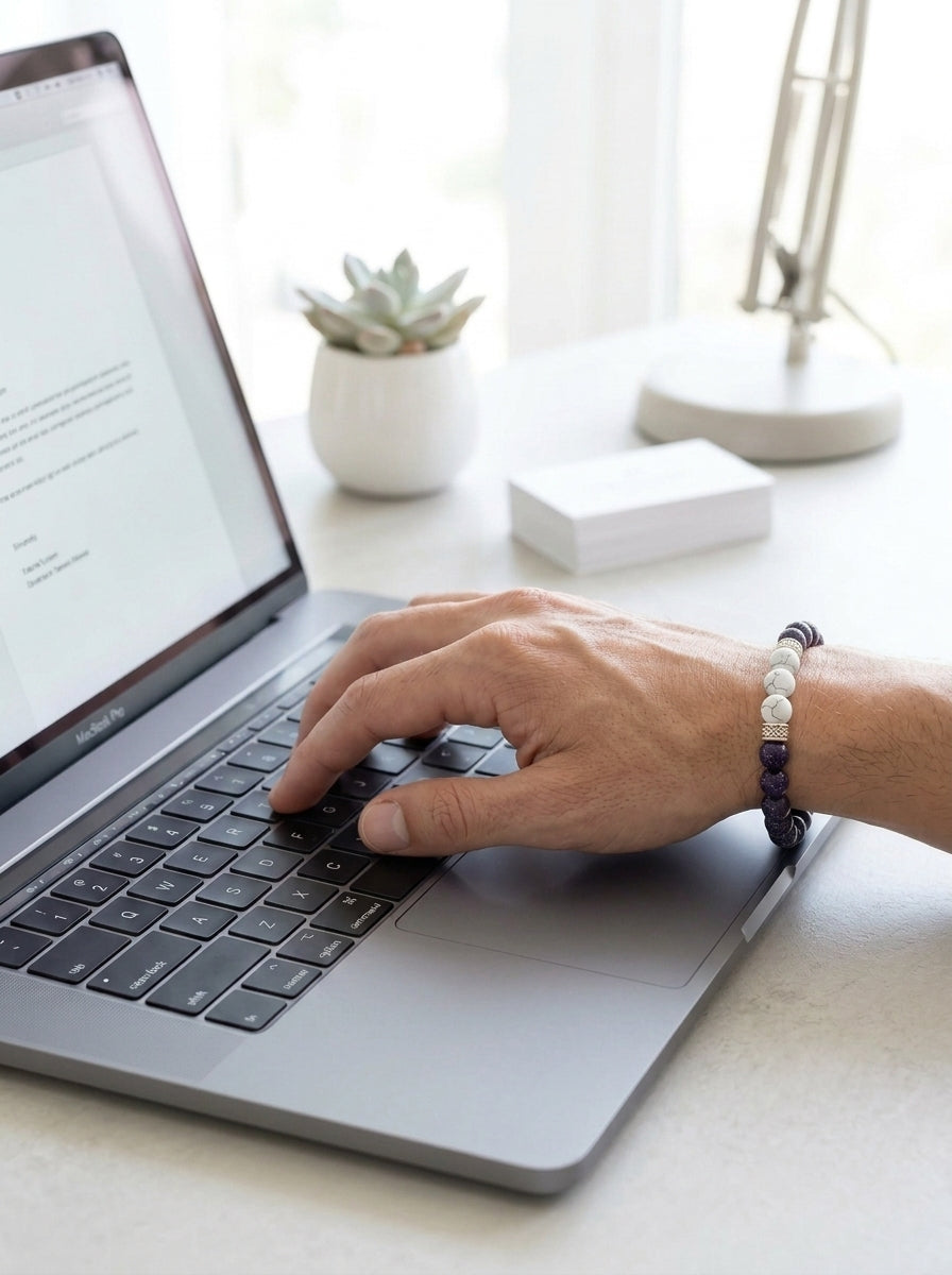A close-up of a person's hand typing on a laptop keyboard, wearing a dark blue and white beaded bracelet. The scene is set on a light-colored desk with a small potted succulent, a white box, and a modern desk lamp in the background.