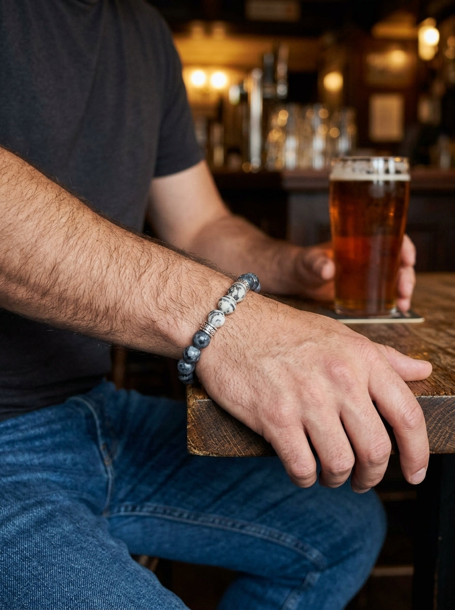 A close-up lifestyle shot of a man at a bar wearing a beaded bracelet on his wrist while holding a glass of amber beer.